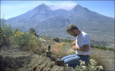 Foto van een vulkanoloog die een seismograaf installeert. Op de achtergrond de vulkaan Mount St. Helens na de eruptie van 18 mei 1980.